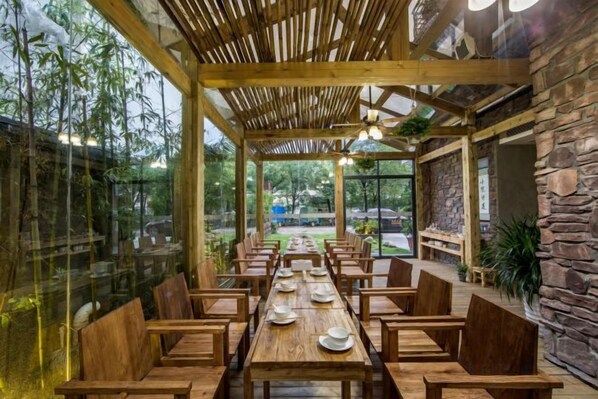 An indoor dining area with wooden tables and chairs, a glass wall, and a view of the outdoors.