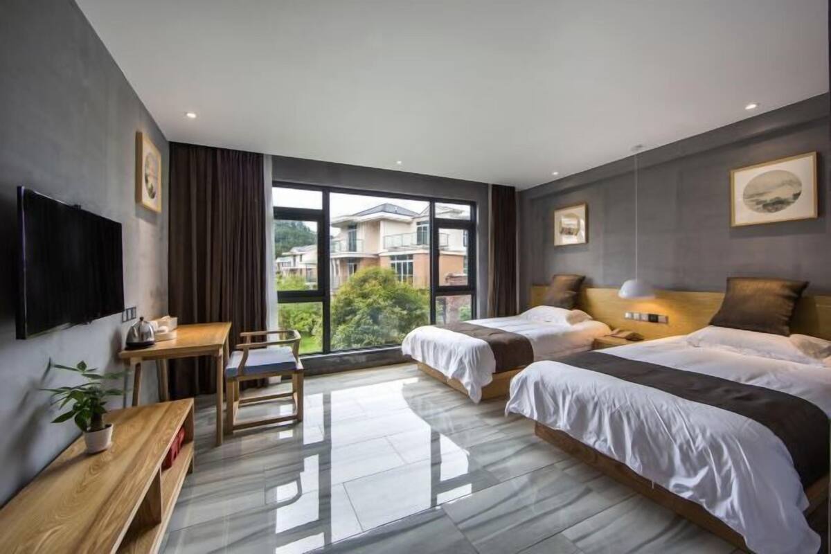 Una habitación de hotel moderna con dos camas, un escritorio y un amplio ventanal con vistas a edificios y zonas verdes.