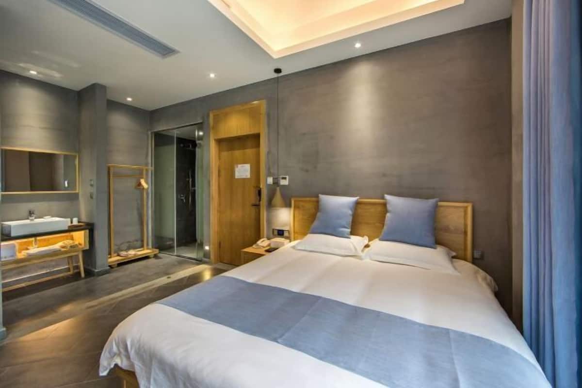 Una habitación de hotel moderna con una cama grande, una mesita de noche, un espejo y un baño con lavamanos y ducha.