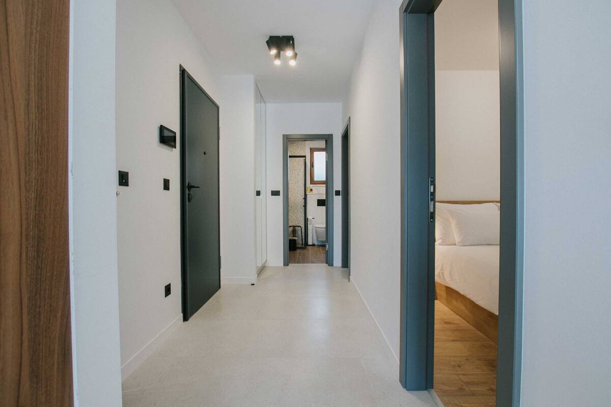 Un pasillo con piso de madera, paredes blancas y una cama a lo lejos.