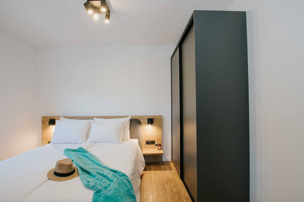 Una habitación de hotel moderna con una cama bien hecha, una mesita de noche con lámpara y un panel grande y oscuro en la pared.