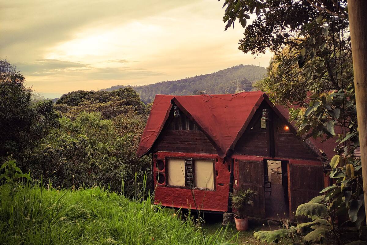 A rustic wooden house with a red roof, surrounded by lush greenery and hills in the background.