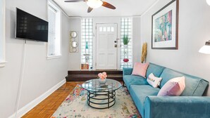 Una sala de estar con un sofá azul, una mesa de centro de vidrio, un ventilador de techo, un televisor y una puerta con paneles de vidrio decorativos.