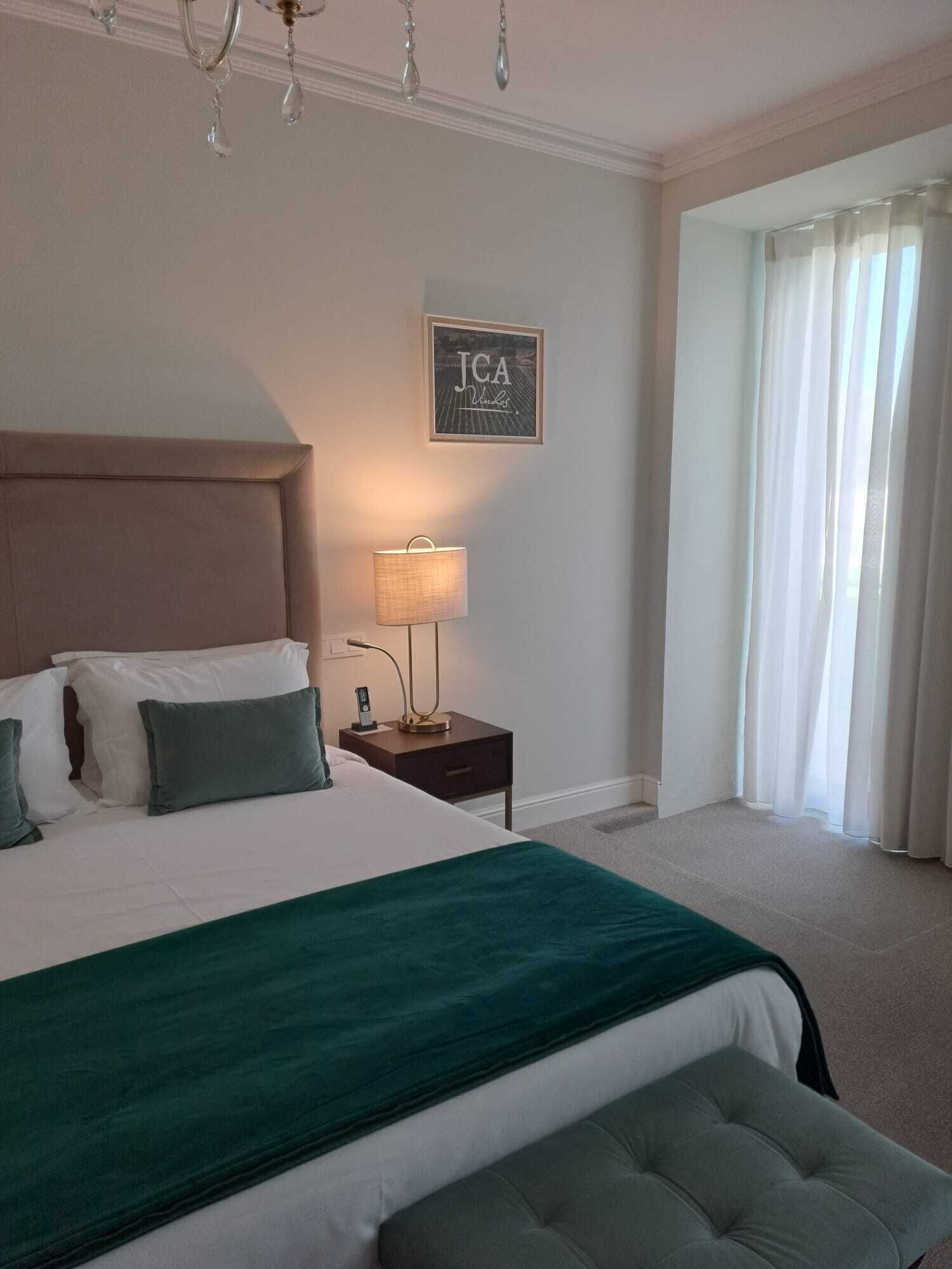 Une chambre d’hôtel avec un grand lit, une table de chevet avec une lampe, un panneau encadré accroché au mur et une fenêtre avec des rideaux.