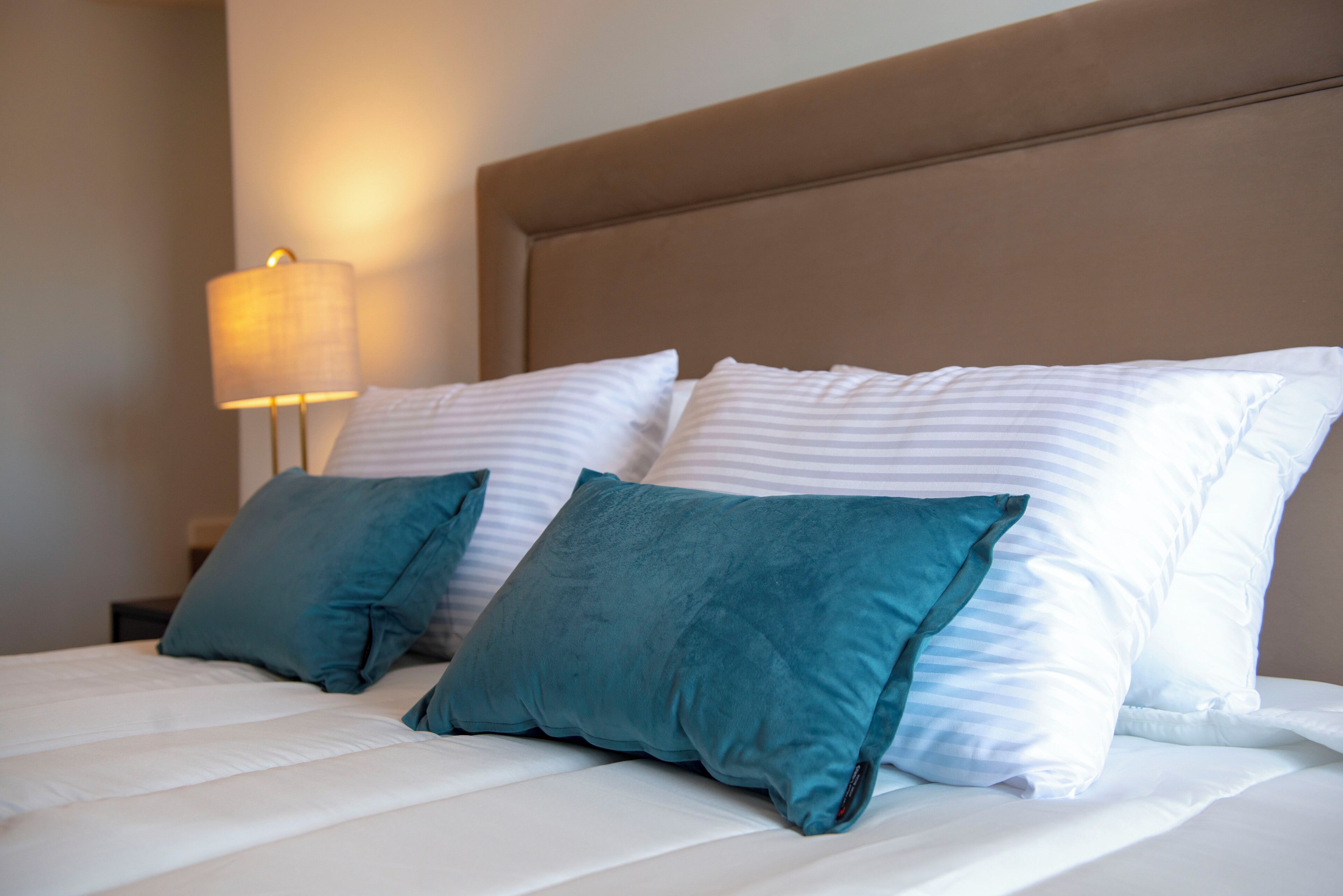 Una cama bien tendida con almohadas blancas y color turquesa, un cabecero color beige y una lámpara de noche.