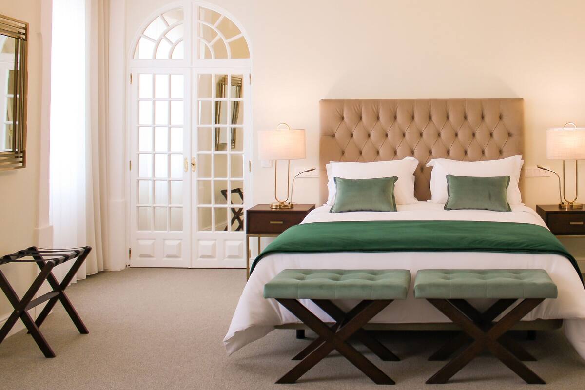 A bedroom with a large bed, two bedside tables, a green bench, and a white door with glass panels.