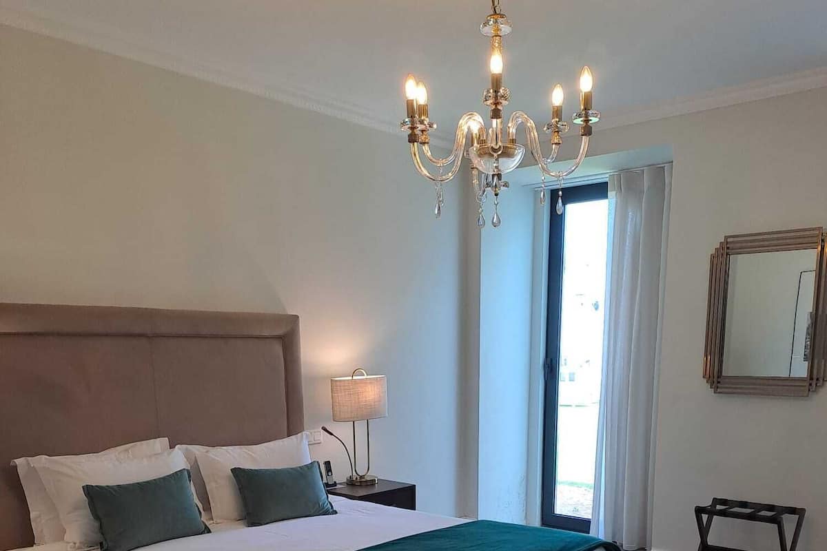 A bedroom with a large bed, a bedside table with a lamp, a bench at the foot of the bed, a chandelier, and a window with curtains.