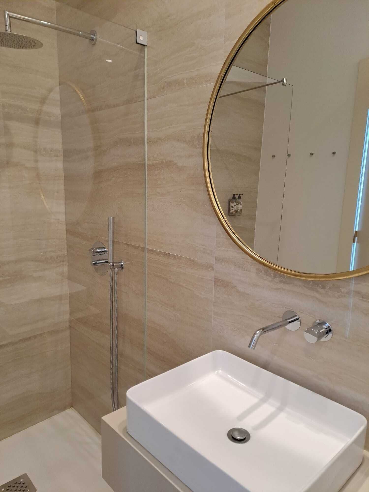 Un baño moderno con ducha envuelta en vidrio, un lavabo blanco y un espejo con marco dorado.