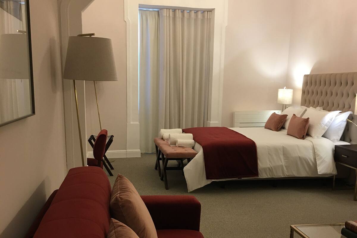 A hotel room with a red sofa, a bed with a red bedspread, a nightstand, a lamp, and a chandelier.