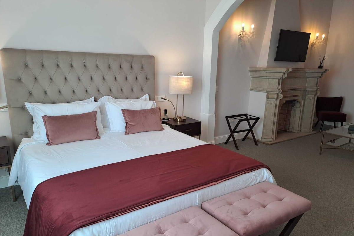 A hotel room with a large bed, a sitting bench, a fireplace, and a TV mounted on the wall.