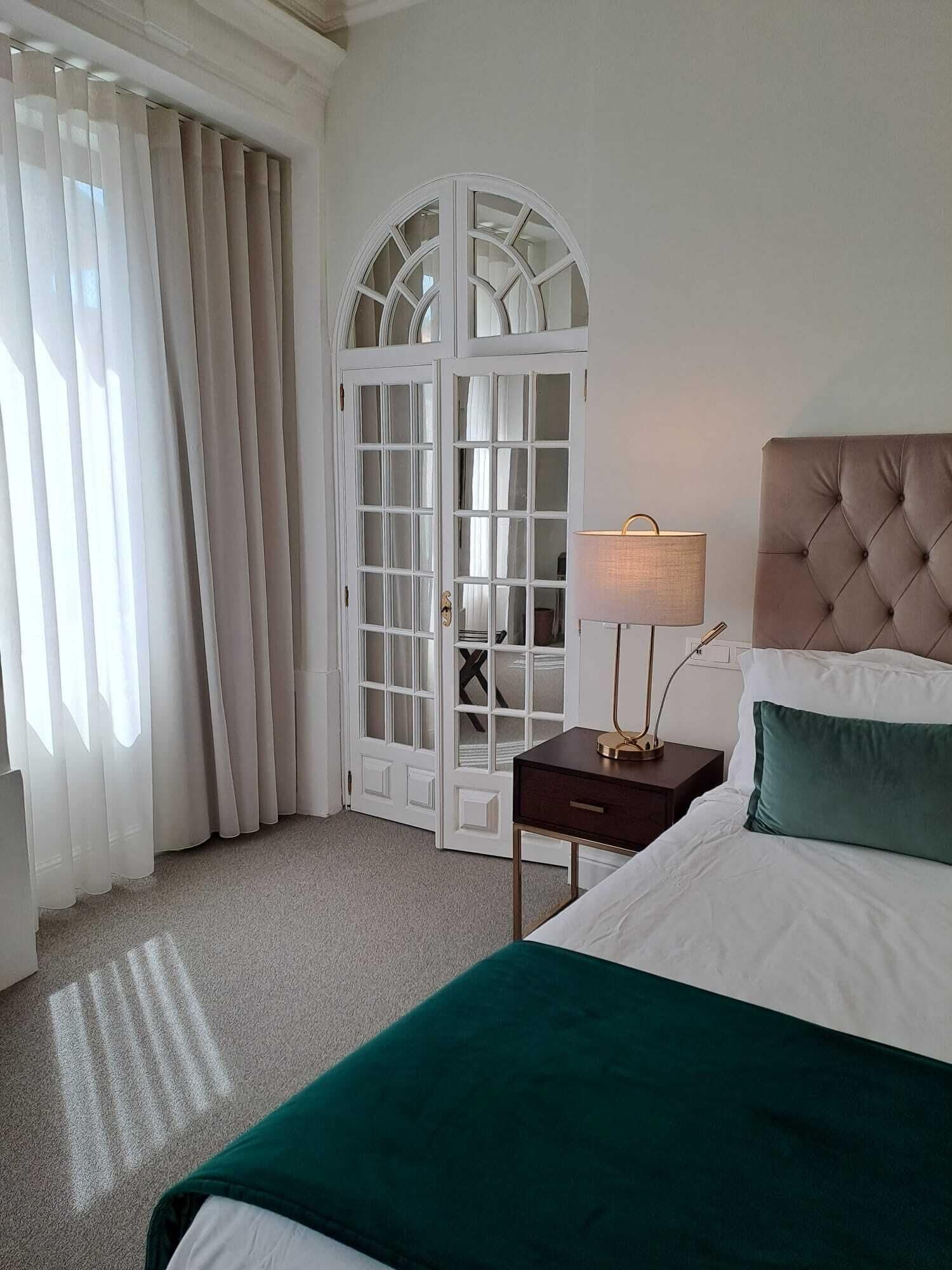 Habitación de hotel con una cama grande, una mesita de noche con lámpara, una ventana con cortinas y una puerta con paneles de vidrio.