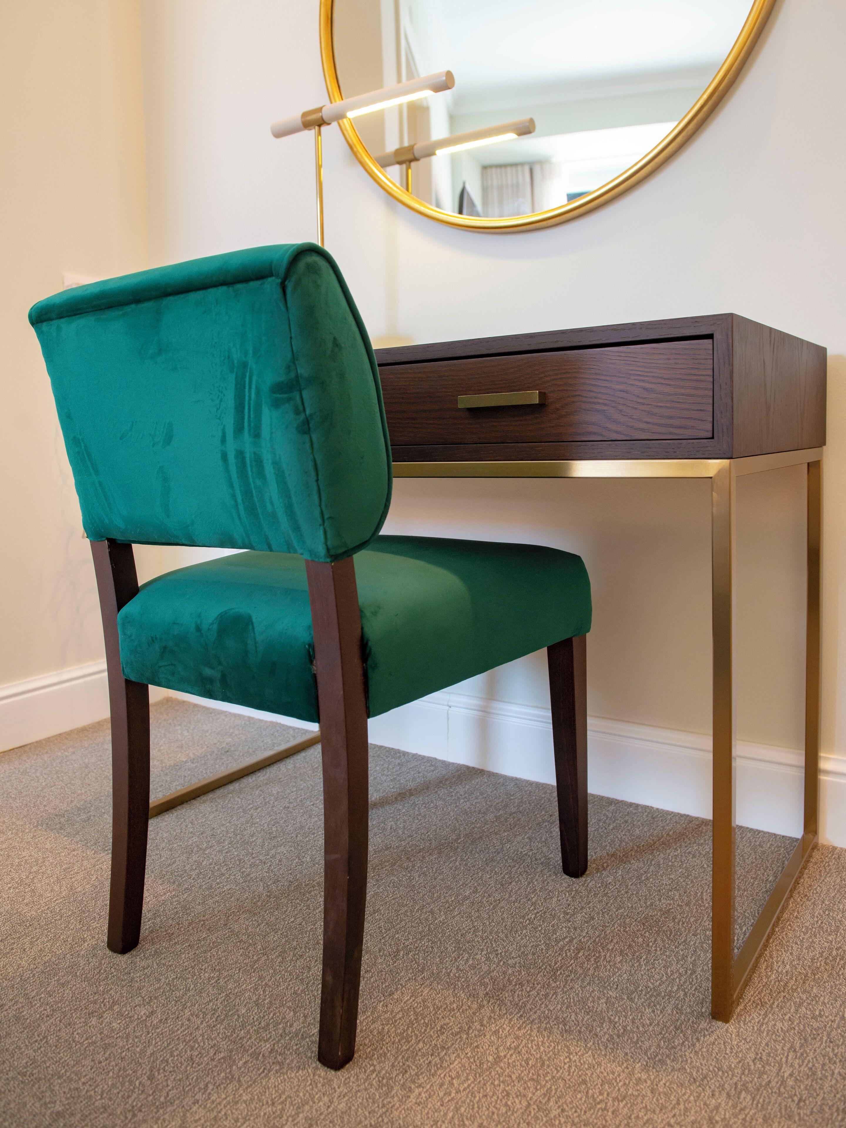 A teal upholstered chair with a wooden frame, a dark wooden desk with a gold accent, and a round mirror with a gold frame.