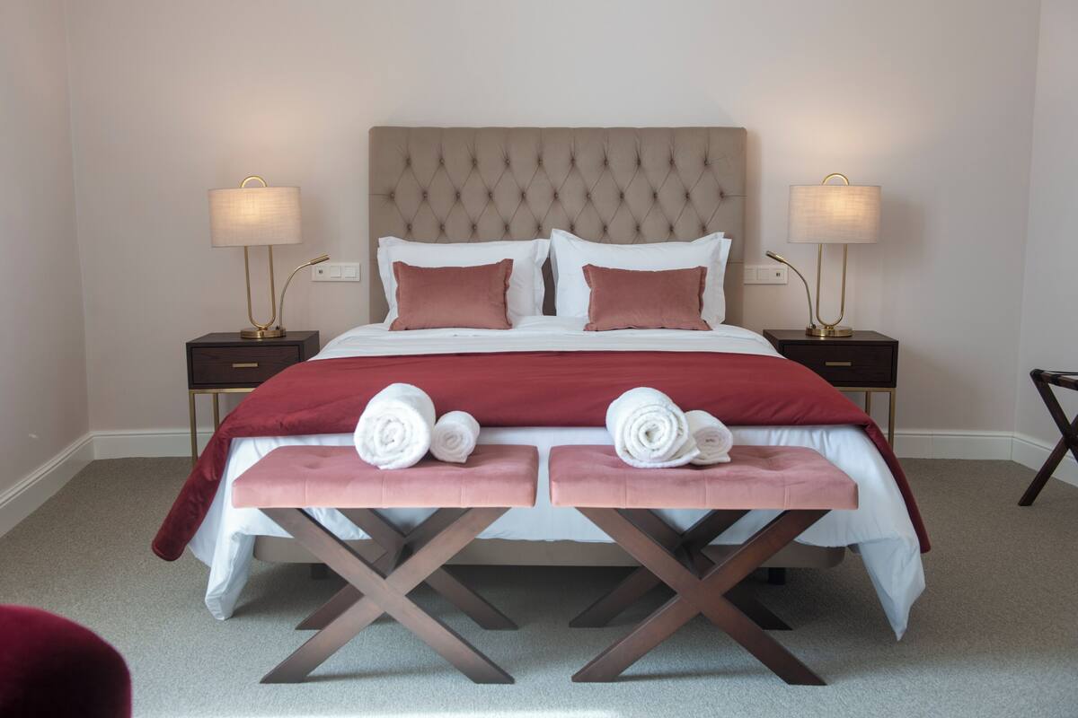A neatly made bed with a tufted headboard, two bedside tables with lamps, and a pair of rolled towels on a bench.