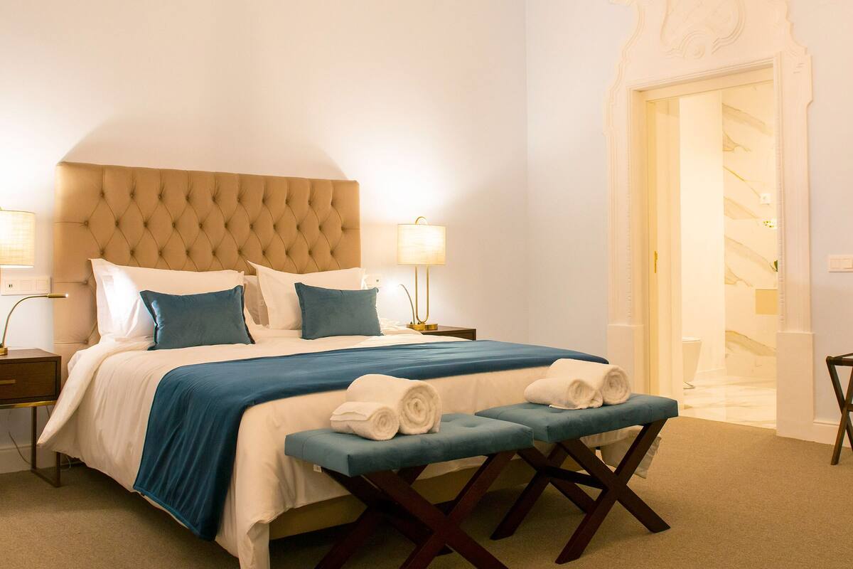 A neatly made bed with a tufted headboard, two bedside lamps, and a bench with towels.