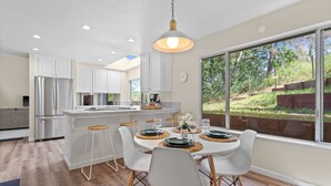 A modern kitchen with white cabinets, a stainless steel refrigerator, a dining area with white chairs, and a large window offering a view of a garden. - Between Napa & San Francisco 6bed 4bath Clean (Concord)