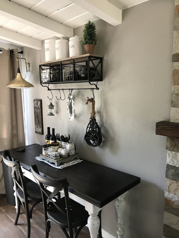 A dining area with a black table, white chairs, a hanging light fixture, and a wall-mounted shelf with kitchen items.
