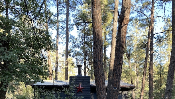 Una cabaña rústica con chimenea de piedra, rodeada de árboles y una escalera de madera que lleva a una terraza.