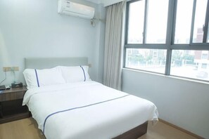 A hotel room with a bed, a nightstand, an air conditioning unit, and a window with curtains.