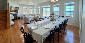 A dining area with a long table set for six, surrounded by chairs. There is a fireplace, a sofa, and a view of the outdoors through large windows.