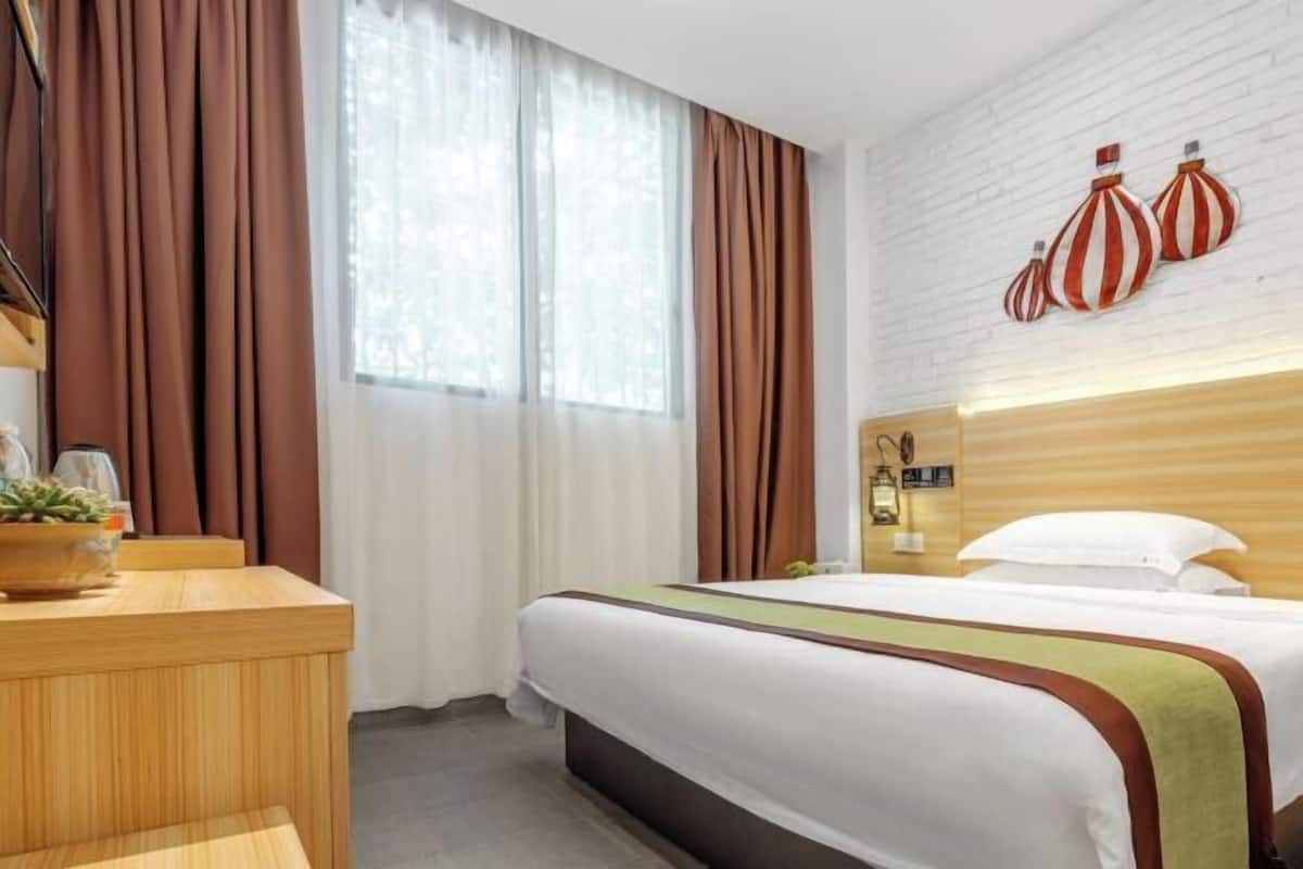 A hotel room with a bed, a wooden desk, a potted plant, and a window with curtains.