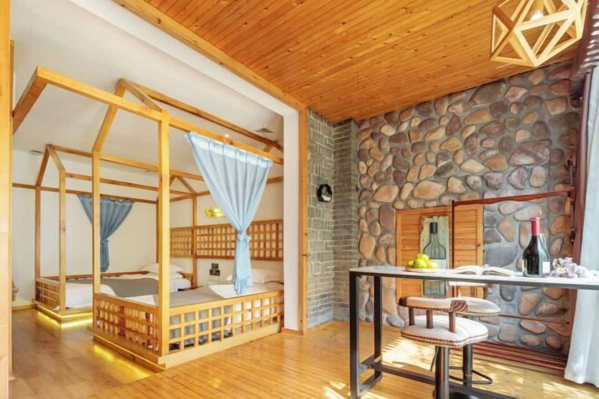 A room with a four-poster bed, stone wall, wooden ceiling, and a table with a bottle and fruit.
