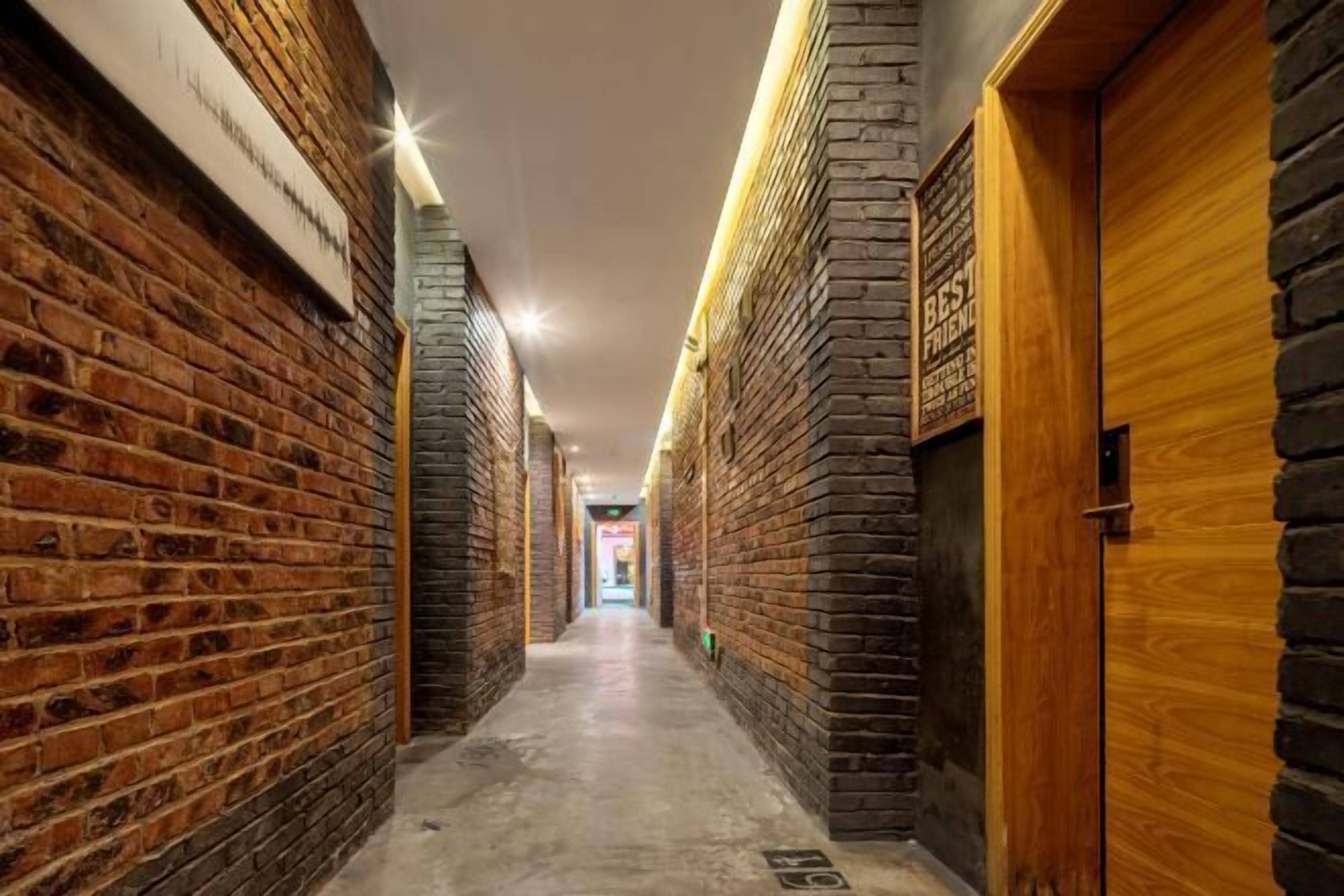 A narrow hallway with brick walls and wooden doors, leading to a brightly lit area at the end.