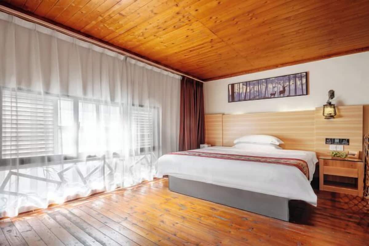 A room with a wooden ceiling, a bed with white bedding, a nightstand, a wall-mounted lamp, and a painting.