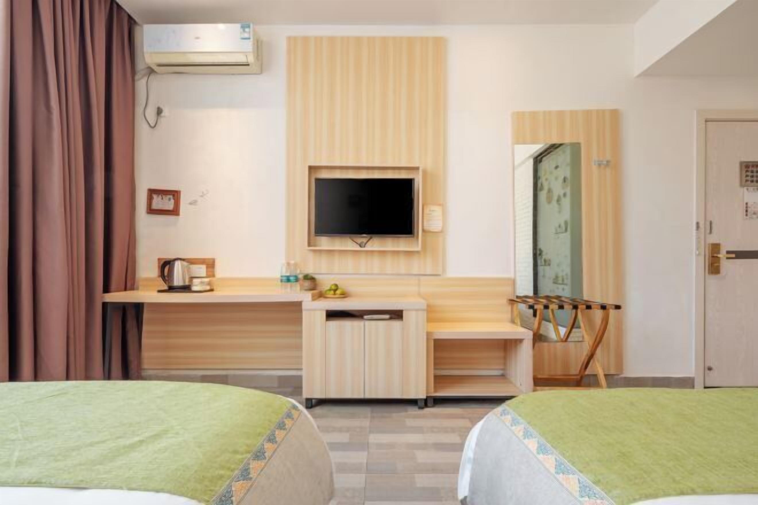 A hotel room with two beds, a TV mounted on the wall, a small desk with a kettle, and a mirror.