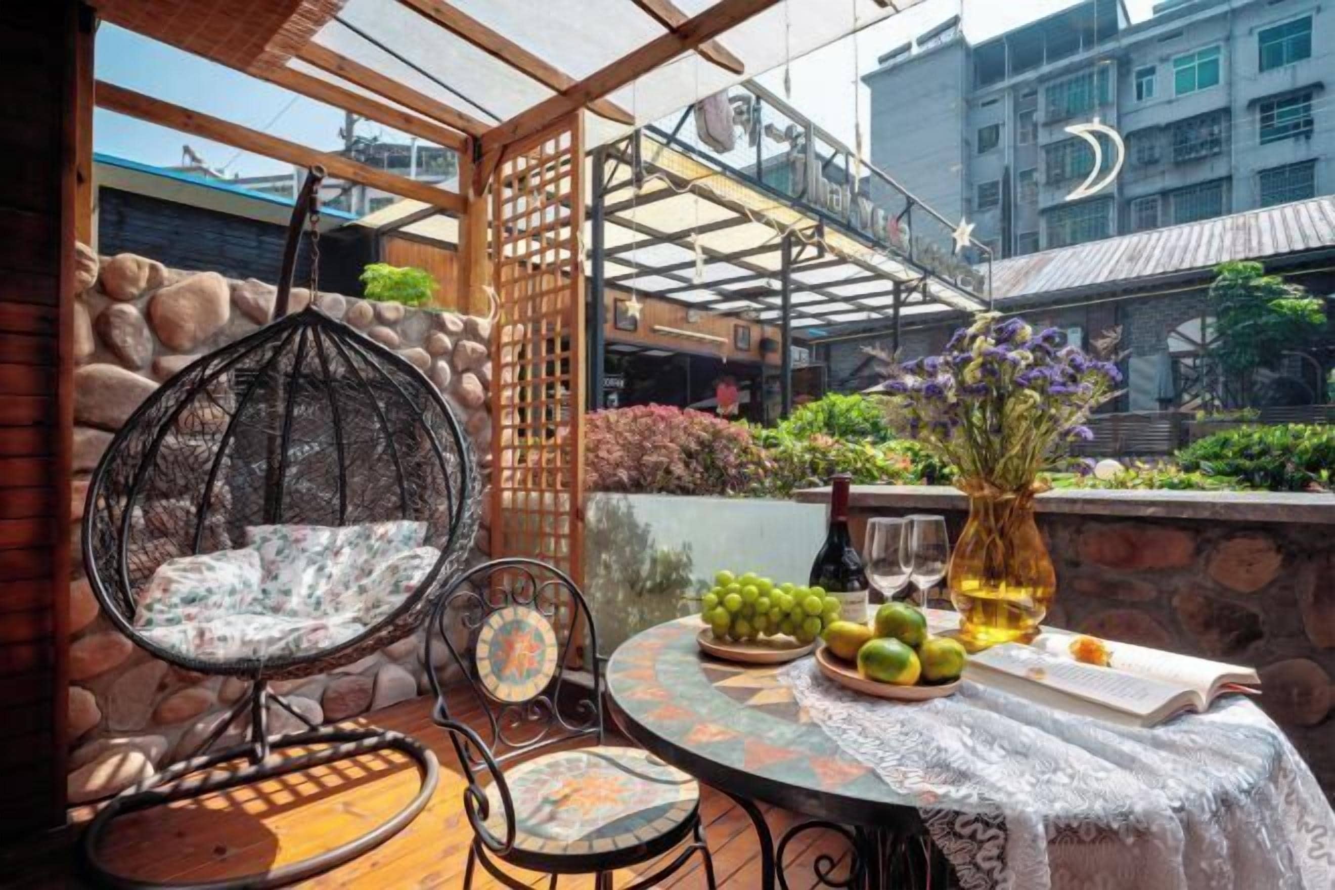 A patio with a hanging chair, a table with fruits and a book, and a view of buildings in the background.