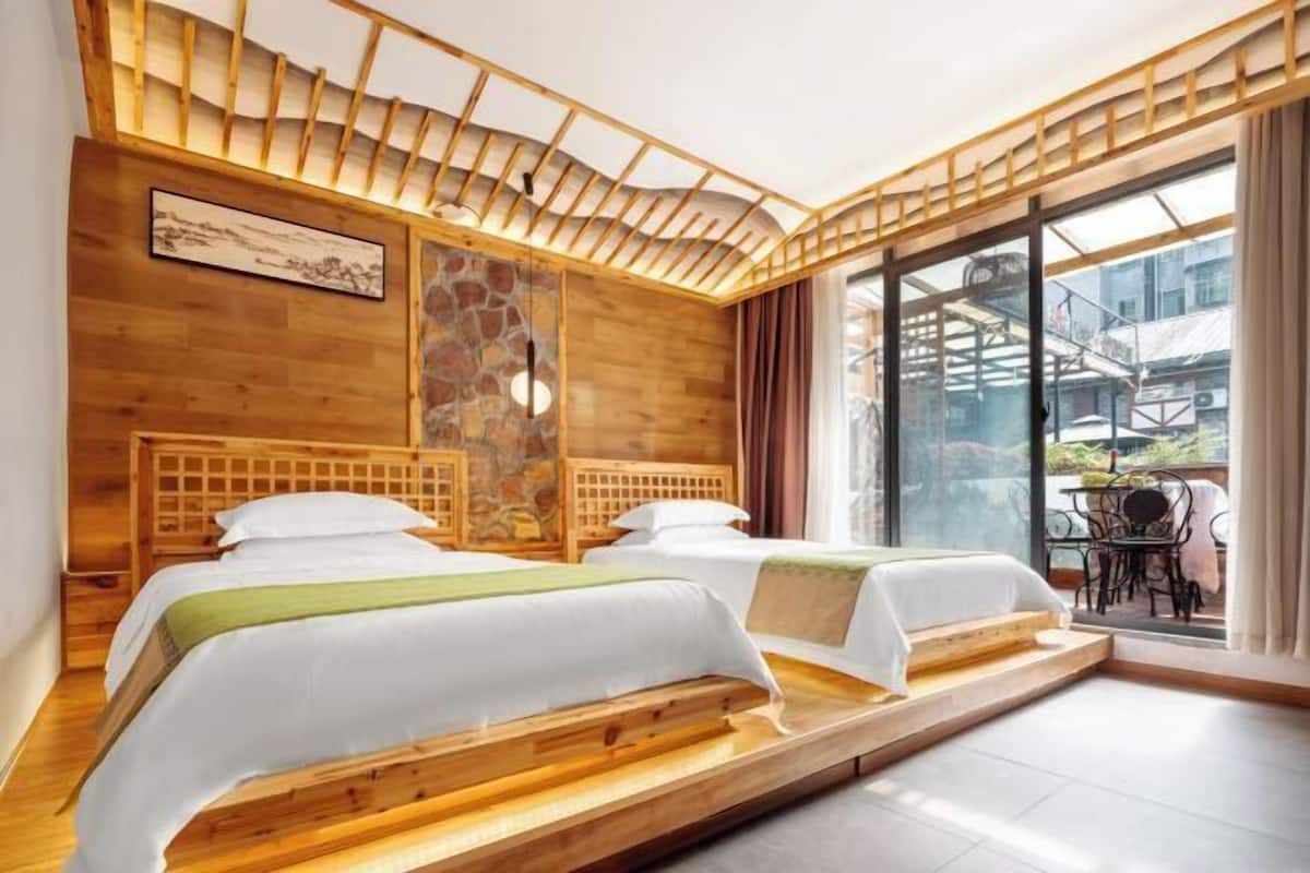 A room with two beds, a stone wall, wooden ceiling beams, and a large window with a view of buildings.