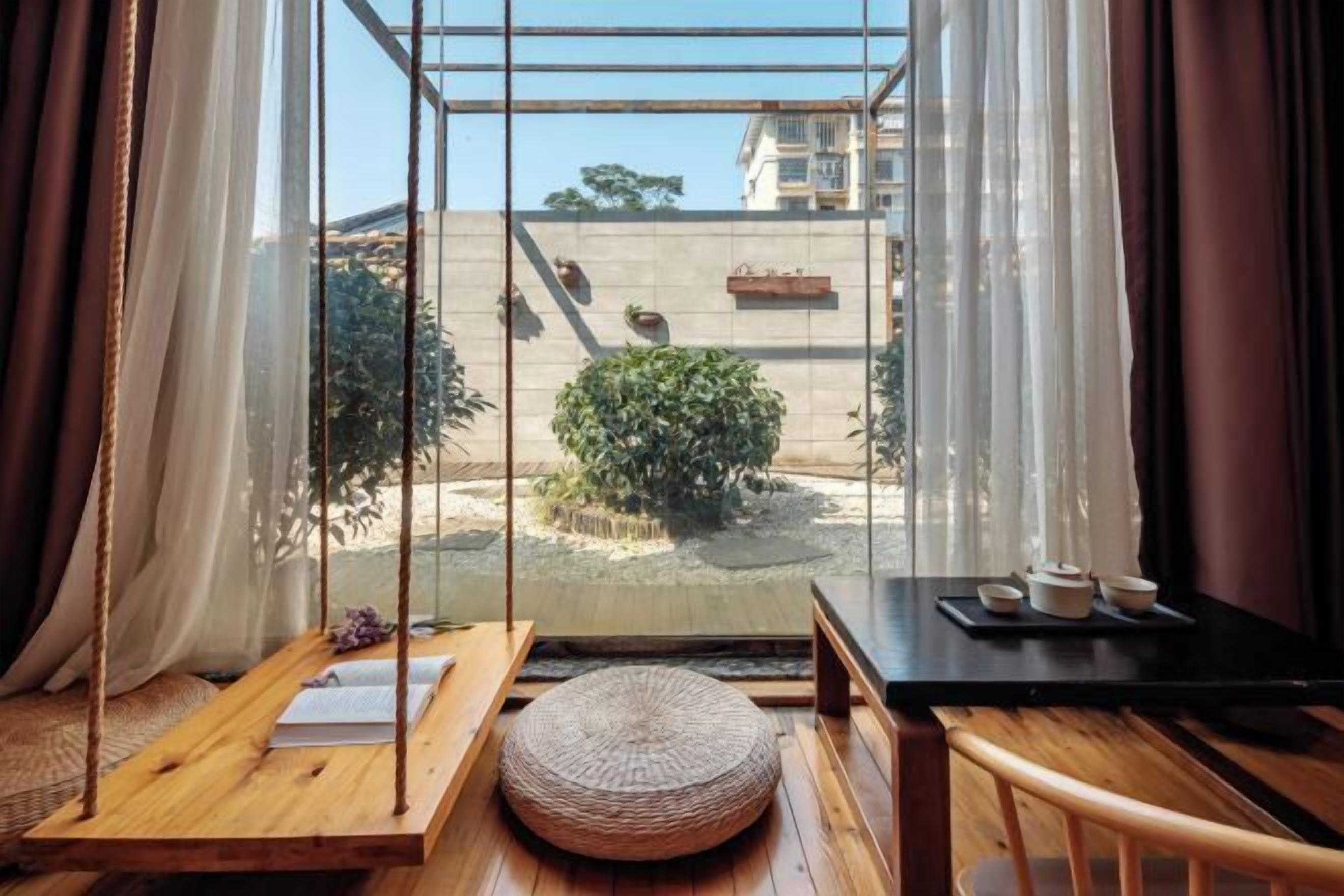A room with a wooden floor, a swing, a round woven ottoman, a table with a black tray, and a view of a garden through a glass window.