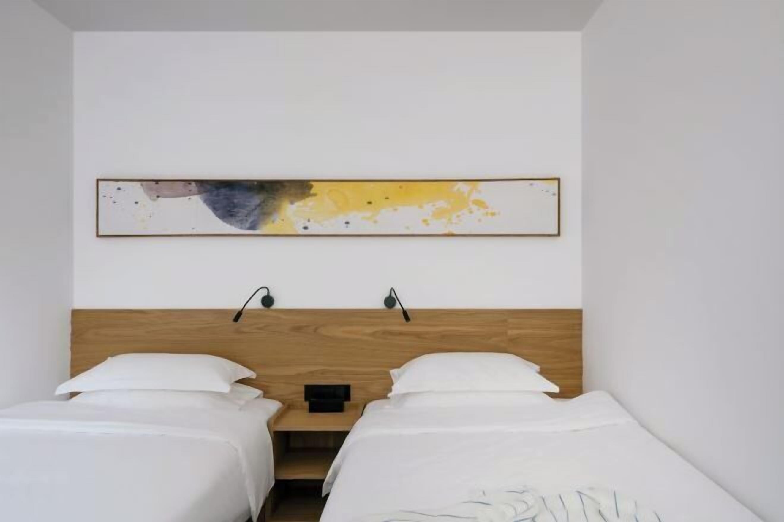 A hotel room with two single beds, a wooden headboard, a wall-mounted abstract painting, and two bedside lamps.