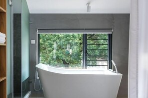 A modern bathroom with a freestanding bathtub, a glass shower enclosure, and a view of greenery outside. - Hanshe Inn (Hangzhou)