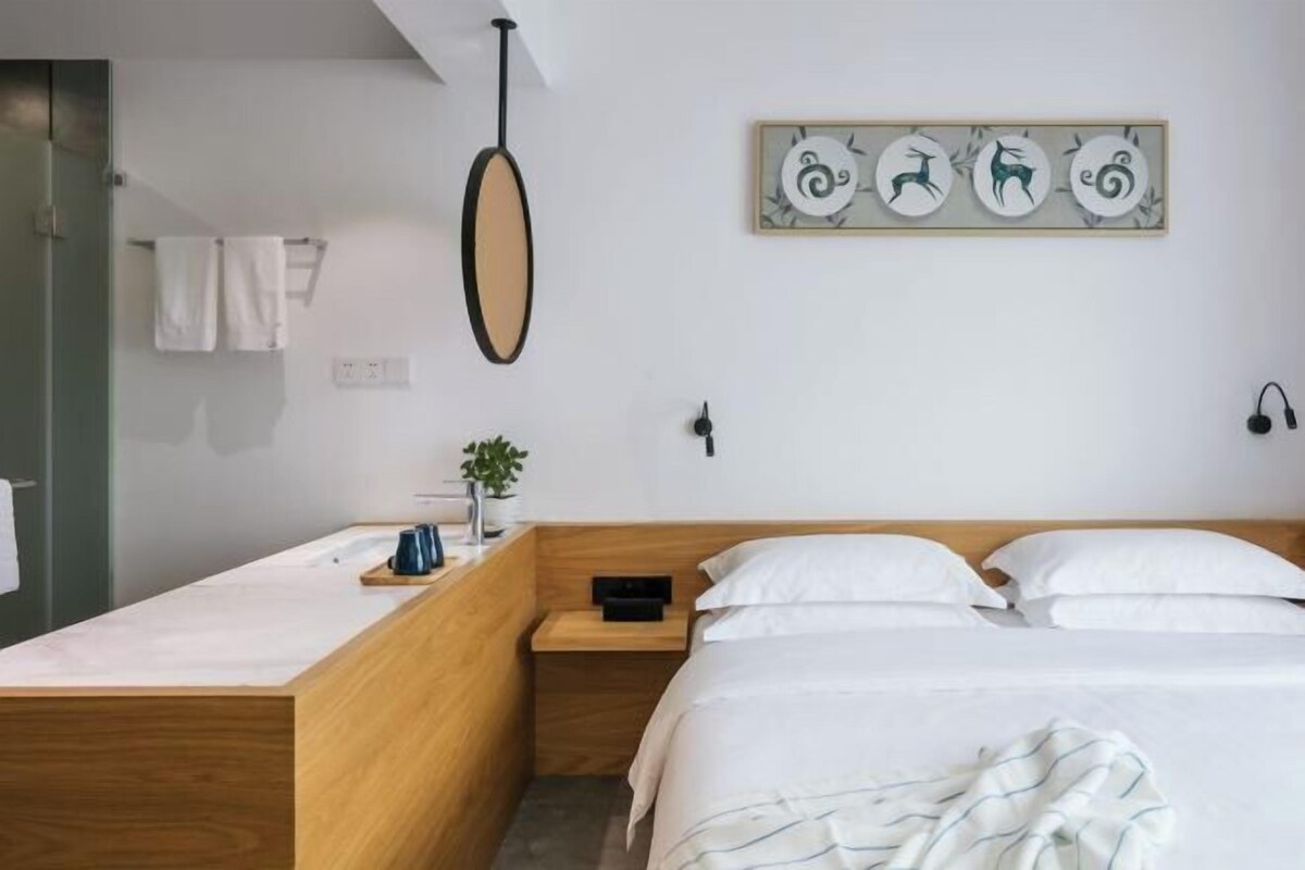 A modern hotel room with a wooden headboard, a bedside table with a plant, a wall-mounted mirror, and a framed artwork above the bed.
