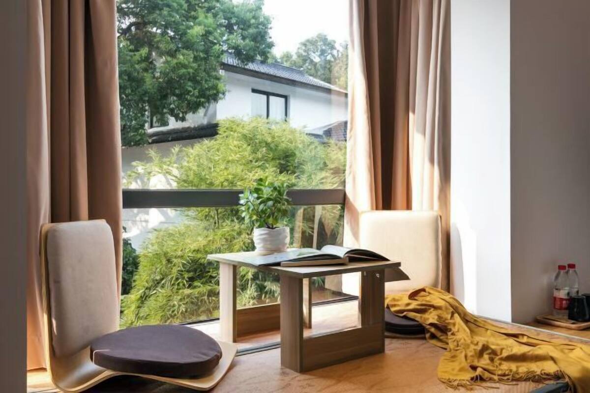 Ein Zimmer mit Blick auf Grünflächen und ein Gebäude. Ein Tisch mit Pflanze und offenem Buch ist zu sehen.