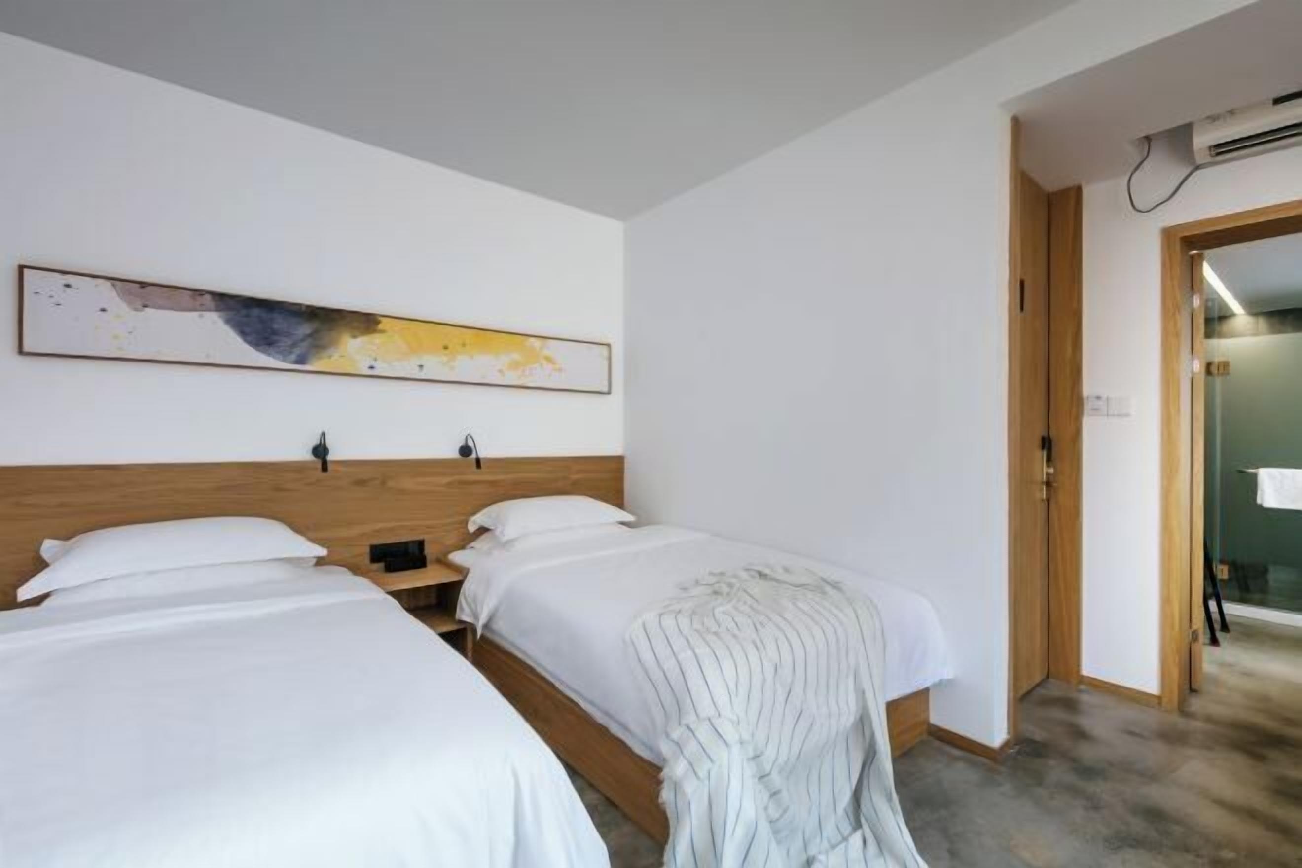A hotel room with two single beds, a wooden headboard, a wall-mounted abstract painting, and a door leading to a bathroom.