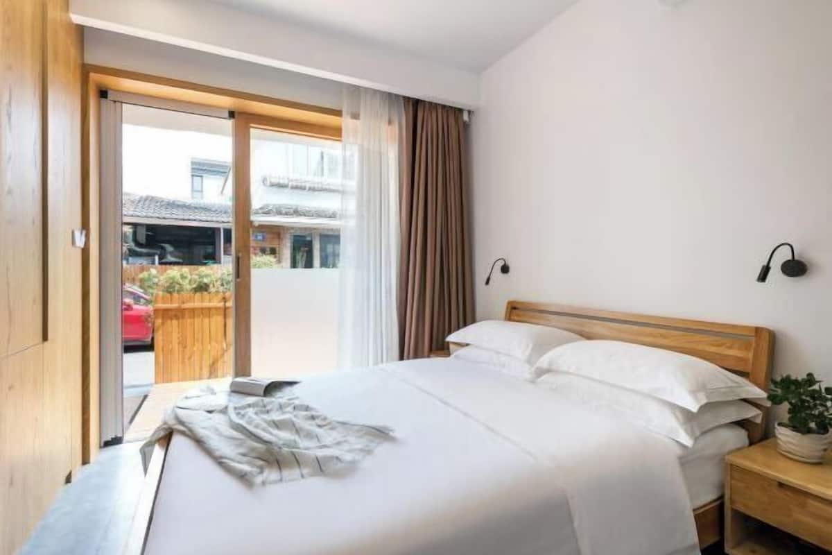 Ein Hotelzimmer mit einem Bett, einem Holzkopfstück, einem Nachttisch, einer Topfpflanze und einer Glastür, die zu einem Außenbereich führt.