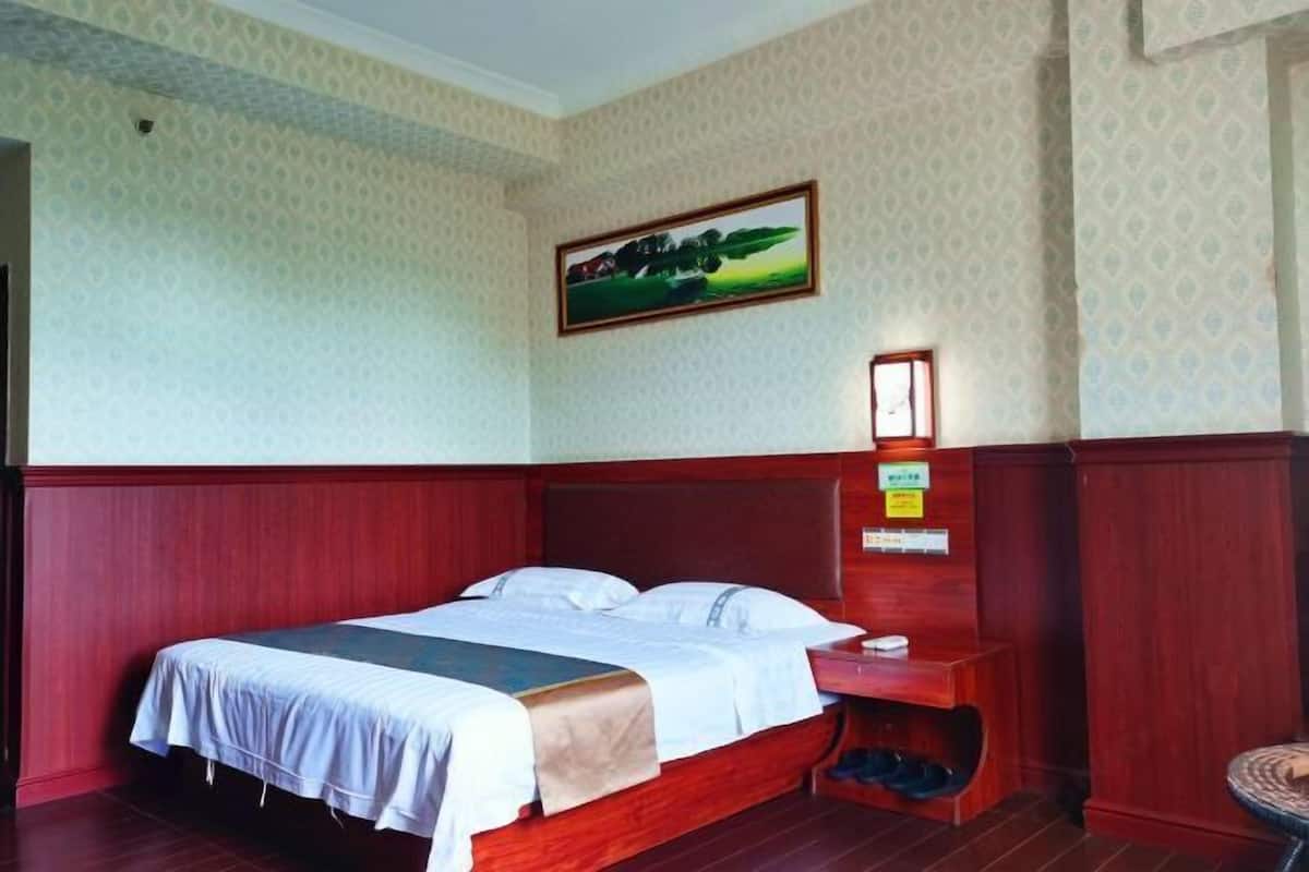 Une chambre d’hôtel avec un lit en bois, une table de chevet, une petite étagère, un tableau encadré au mur et une fenêtre.