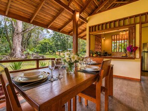 A wooden dining table set with plates, glasses, and a vase of flowers, overlooking a lush garden and a kitchen area.