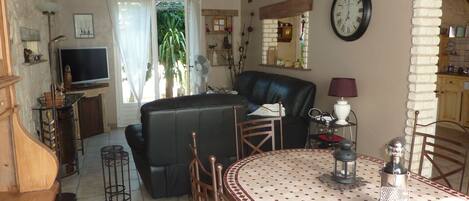 Una sala de estar con un sofá de cuero negro, una mesa de comedor con mantel estampado, un juego de comedor de madera, un televisor y un reloj en la pared.