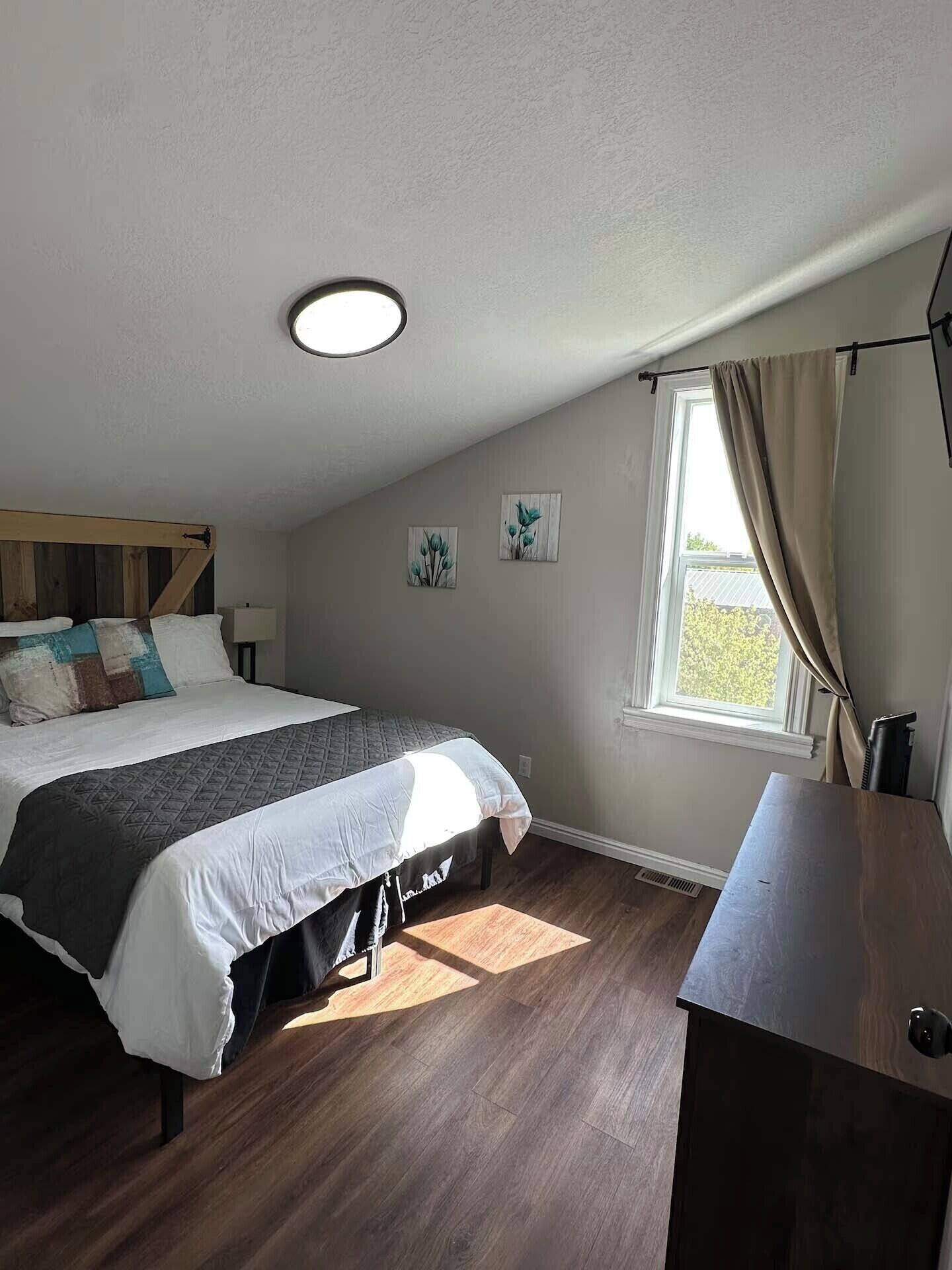 A bedroom with a bed, a desk, a window with curtains, and a ceiling light.