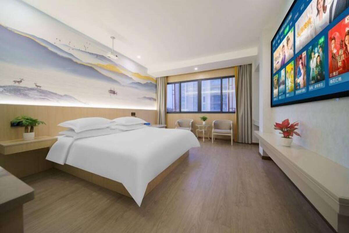 A hotel room with a bed, a desk, a chair, a small table, a wall-mounted TV, and a mural on the ceiling.