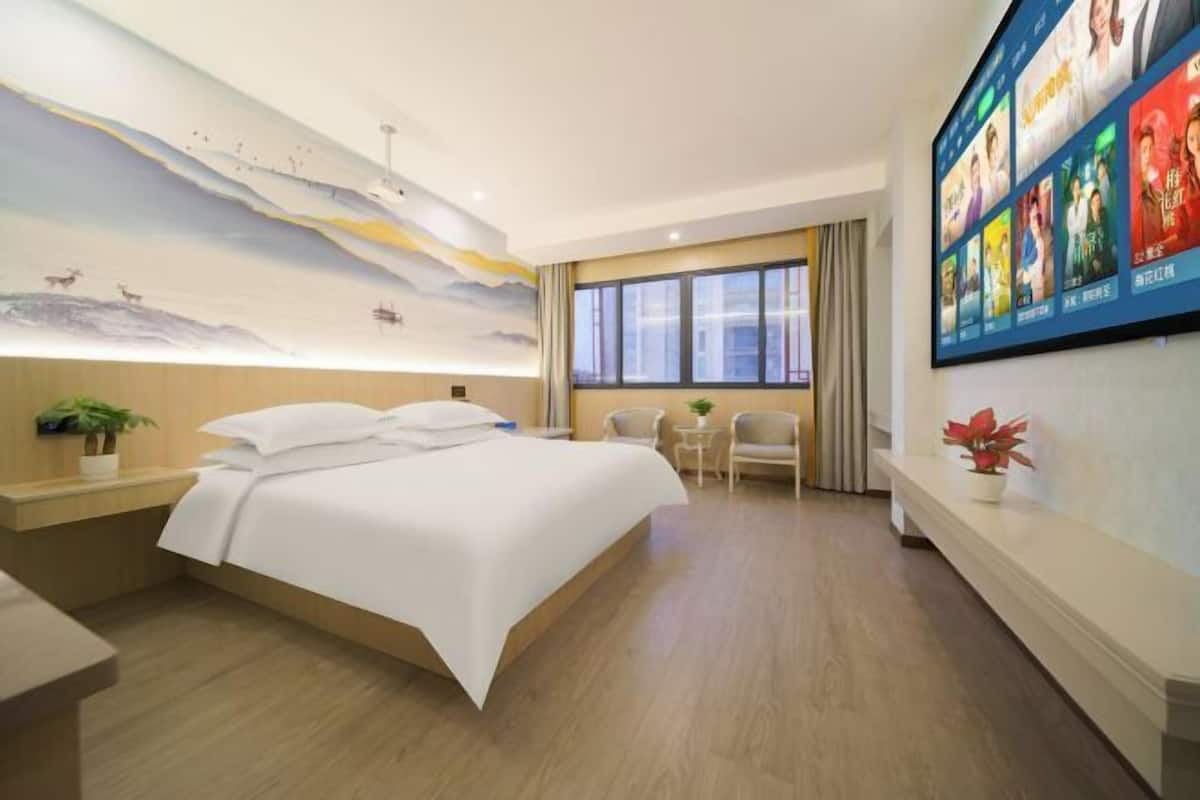 A hotel room with a large bed, a mural on the wall, a TV screen displaying movie posters, and a small table with chairs.