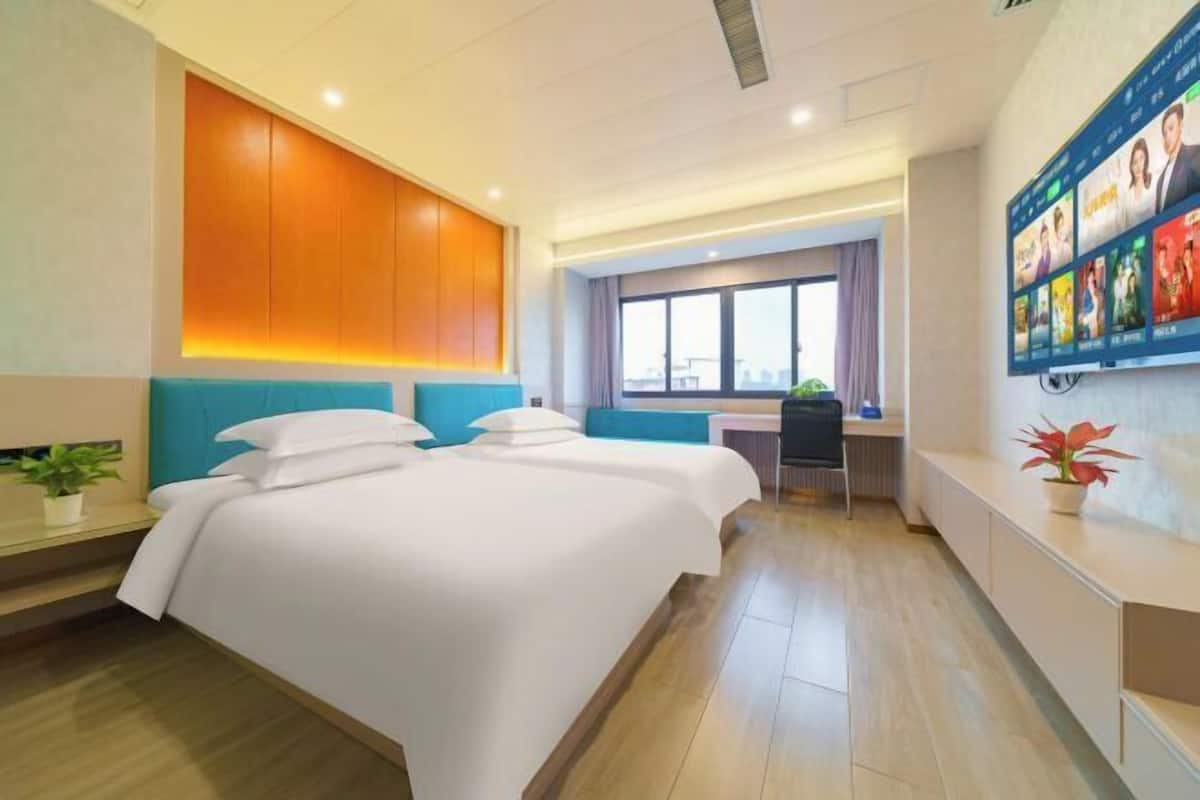 A hotel room with two beds, a desk, a window with a city view, and a wall-mounted TV.