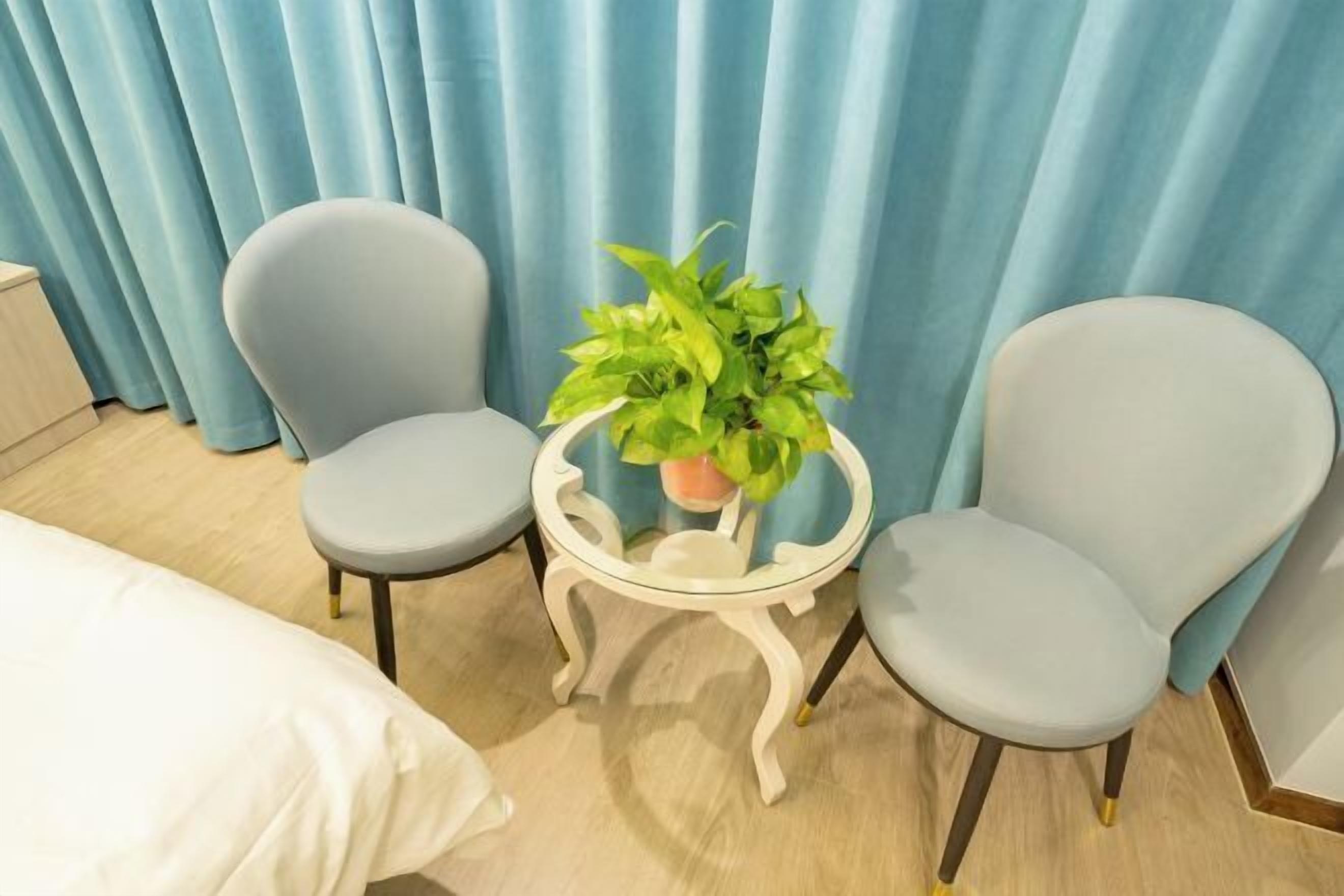 Um quarto com duas poltronas cinzas, uma mesinha branca com uma planta e uma cama com roupa de cama branca.