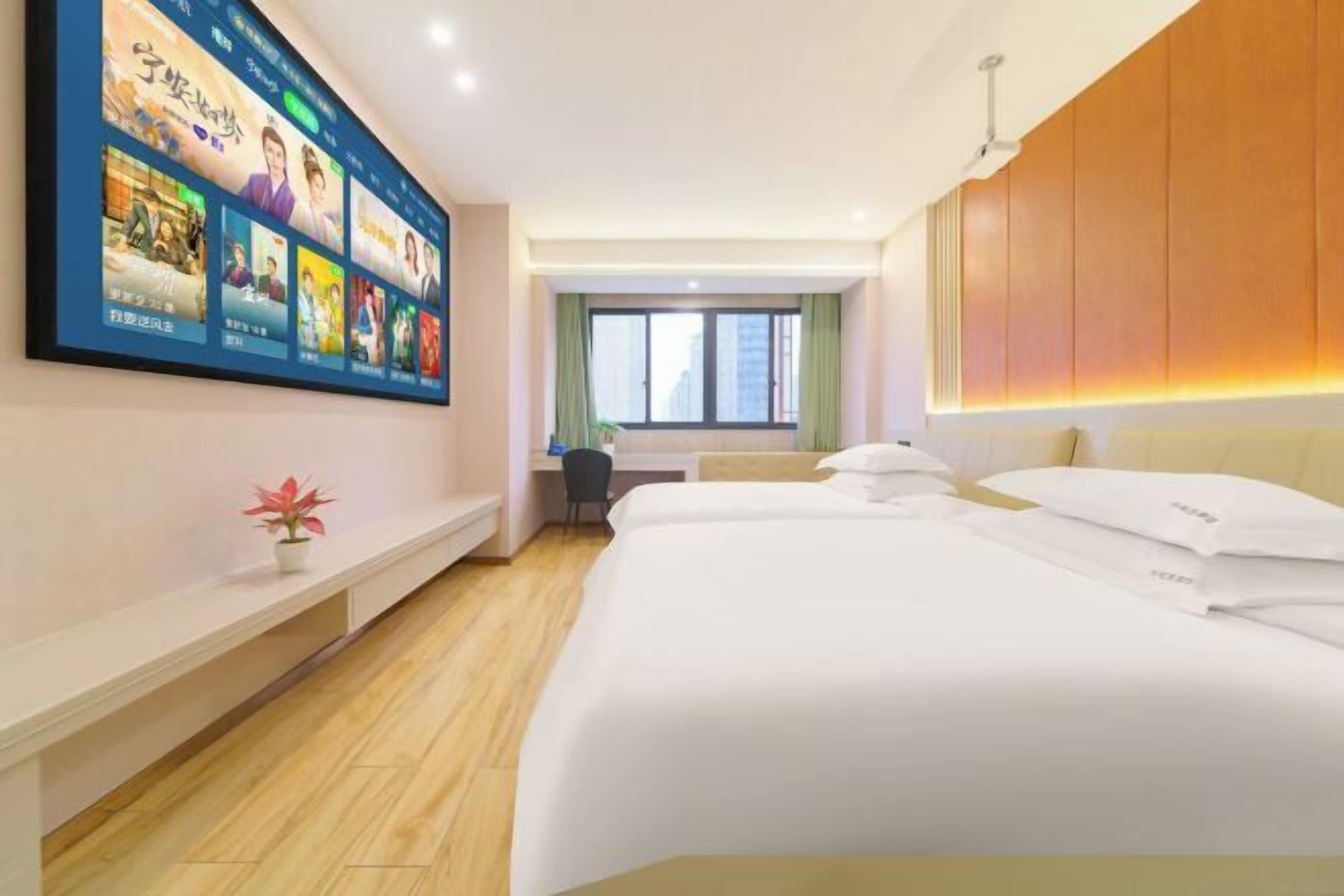 Habitación de hotel con una televisión grande mostrando carteles de películas, un escritorio con una silla, una cama con ropa de cama blanca y una ventana con cortinas.