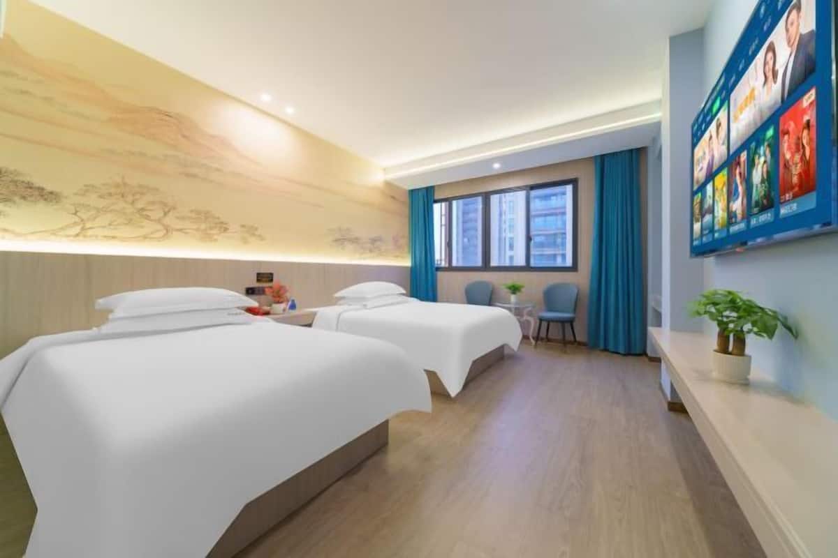 A hotel room with two beds, a small table, a chair, a TV, and a wall with framed pictures.