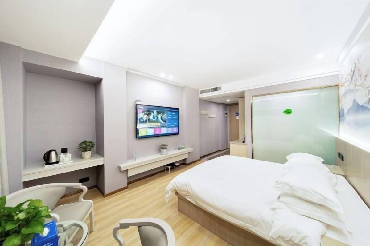A modern hotel room with a large bed, a flat-screen TV mounted on the wall, a small desk with a chair, and a plant.