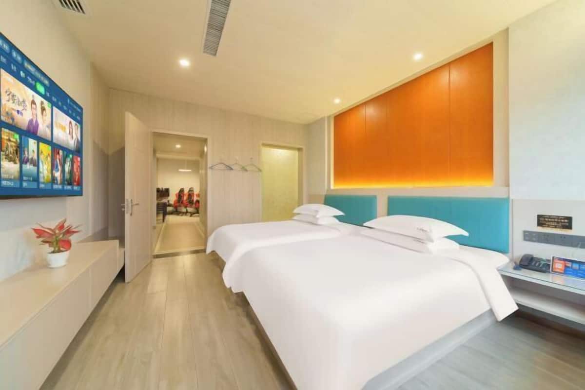 A hotel room with two beds, a TV wall, a plant, and a desk with a phone.