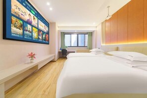 Habitación de hotel con un televisor de pared grande mostrando varios programas, una cama con ropa blanca, un escritorio con una silla y una ventana con cortinas.